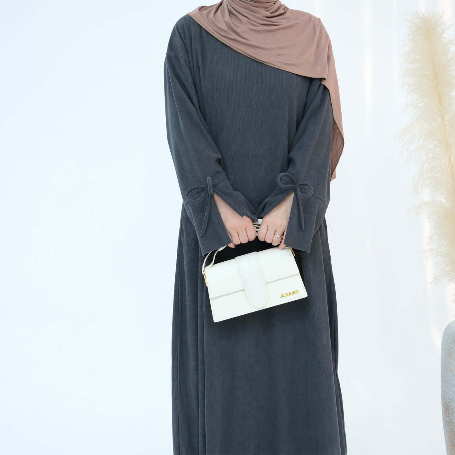 Nawal Corduroy Abaya in dark gray with adjustable ties at sleeves and a stylish white handbag.